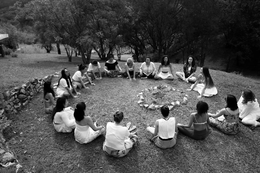 A group of diverse women sits in a circle around a fire pit in a grassy meadow.