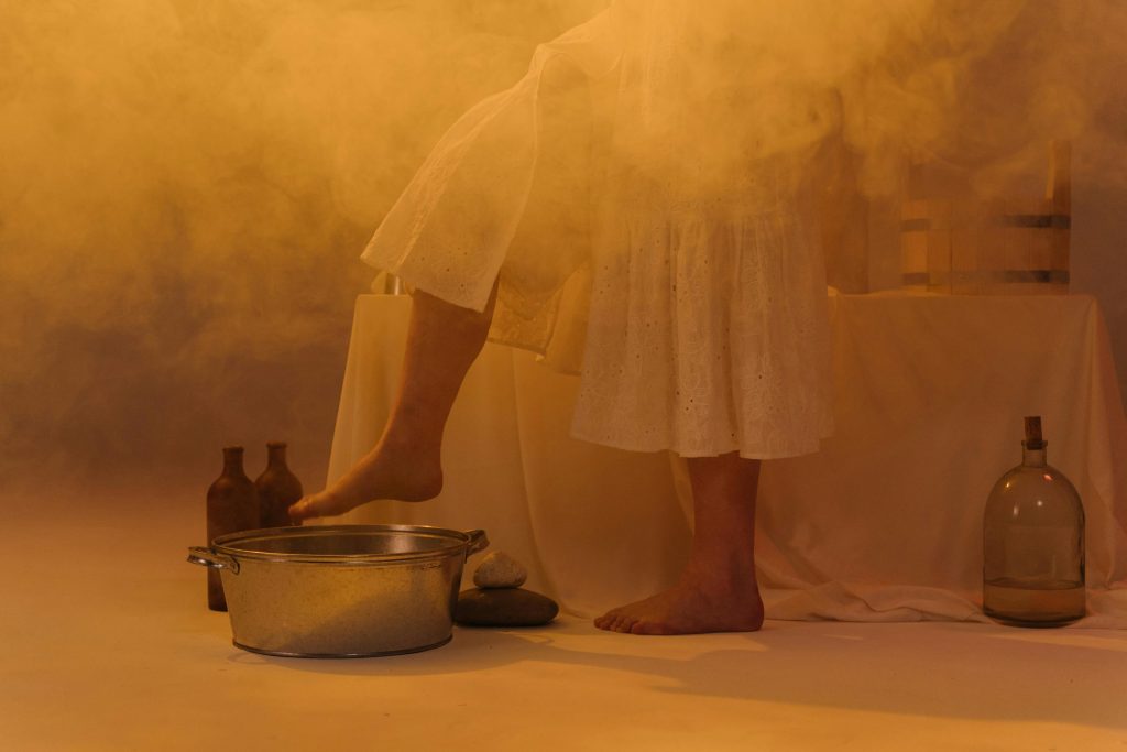A serene setting featuring a woman soaking her feet in a metal basin with glass bottles and smoke.